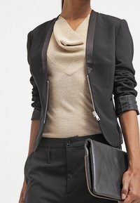 Kobieta w czarnej marynarce z podwiniętymi rękawami, beżowej bluzce z golfem, czarnych spodniach, trzymająca czarną skórzaną kopertówkę z zamkiem błyskawicznym.