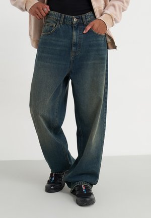 Personne portant un jean bleu foncé ample, des baskets noires, un haut noir et une veste beige, les mains dans les poches avant, debout sur un sol clair.