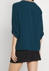 Maglietta teal a tre quarti in tessuto leggero, con un fit comodo e una texture sottile. Indossata con una gonna nera.