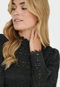 Juoda nėrinių palaidinė su reljefiniu raštu, aukšta apykakle ir raukiniais dekoruotu nėrinių apvadu ant apykaklės ir rankogalių. Matomas minkštas, sudėtingas raštas.