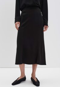 Femme portant un haut noir à manches longues, une jupe midi noire avec des détails de couture, et des ballerines noires à bouts pointus avec des œillets en argent, se tenant sur un sol blanc.