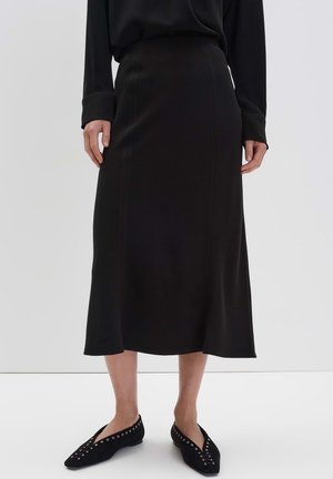 Frau in einem schwarzen Langarmtop, schwarzer Midi-Rock mit Nahtdetails und schwarzen spitzen Ballerinas mit silbernen Ösen, stehend auf einem weißen Boden.