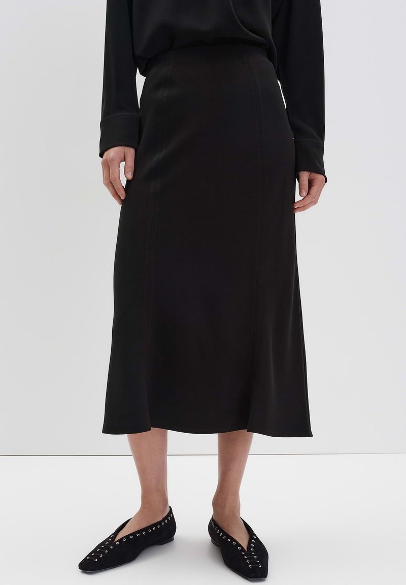 Femme portant un haut noir à manches longues, une jupe midi noire avec des détails de couture, et des ballerines noires à bouts pointus avec des œillets en argent, se tenant sur un sol blanc.