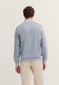 Hellblauer Pullover mit strukturiertem Gewebe, geripptem Saum und Bündchen sowie kontrastierenden Schulterakzenten. Getragen mit khakifarbenen Hosen. Rückansicht.