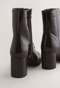 Sorte laklæder ankelstøvler med et strømlinet design, der har lynlås i siden, blokhæl og glat tekstur. Tykke syningsdetaljer er synlige.