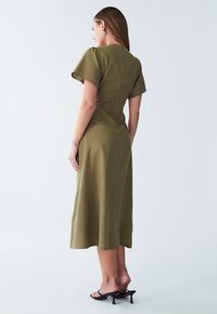 Ženska v olivno zeleni midi obleki s kratkimi rokavi in črnimi petami, stoji obrnjena stran od kamere na enobarvni beli podlagi.