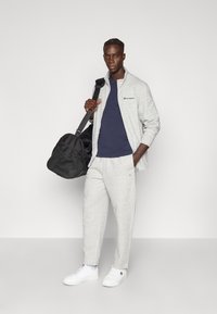 Ανοιχτό γκρι ζακέτα με φερμουάρ πάνω από μπλε σκούρο πουκάμισο, συνδυασμένο με αντίστοιχο παντελόνι φόρμας και λευκά αθλητικά παπούτσια. Μια μαύρη τσάντα duffle κρατιέται στο δεξί χέρι.