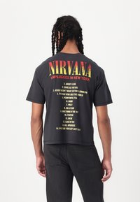 Černé tričko s nápisem "NIRVANA UNPLUGGED IN NEW YORK" v žluté a červené barvě, na zadní straně s vytištěným seznamem názvů písní v žluté barvě.