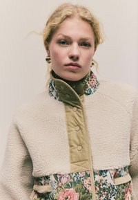 Ung kvinde med blond hår, iført en blomsterpræget jakke med beige fleece og guld hoop øreringe mod en ensfarvet baggrund.