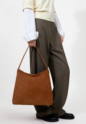 Persoon met een crème trui en wit overhemd houdt een grote bruine suède handtas met dunne handvatten vast, gecombineerd met losse donkere broek en zwarte schoenen.