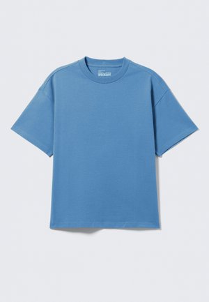 T-shirt uni à manches courtes et col rond en bleu pastel, posé à plat sur un fond gris clair, avec étiquette de la marque visible à l'intérieur du col.