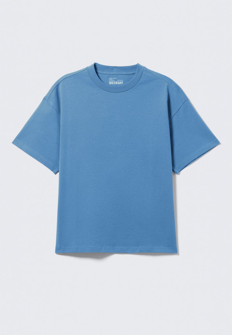 T-shirt uni à manches courtes et col rond en bleu pastel, posé à plat sur un fond gris clair, avec étiquette de la marque visible à l'intérieur du col.