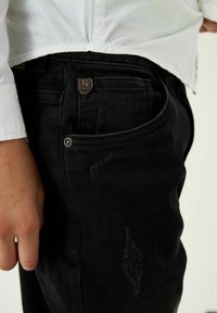 Vaqueros de mezclilla negra con corte ajustado, que presentan un parche de cuero con el logo, detalles sutiles de desgaste y bolsillos estándar.