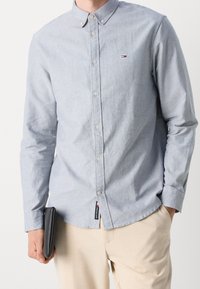 Muž v světle modré košili s dlouhým rukávem a béžových kalhotách, držící černou peněženku v levé ruce ve výšce boků.