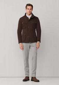 Mand iført en mørkebrune knapjakke med fire forlommer, lysegrå bukser og brune ruskind støvler, stående imod en ensfarvet baggrund.