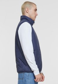 Navy quilted vest with high collar, front zipper, and side pockets; worn over a long-sleeve white shirt, displayed on a neutral background.