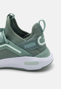 Zapatilla deportiva de malla verde con parte superior texturizada, acentos tonales y una mediasuela blanca. Cuenta con una correa con marca y una lengüeta en el talón.