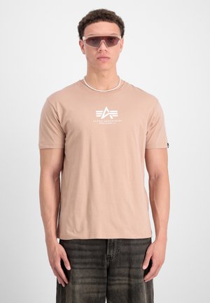 Camiseta de color rosa claro con un diseño de logo blanco, de manga corta y cuello redondo, combinada con jeans oscuros y gafas de sol claras.