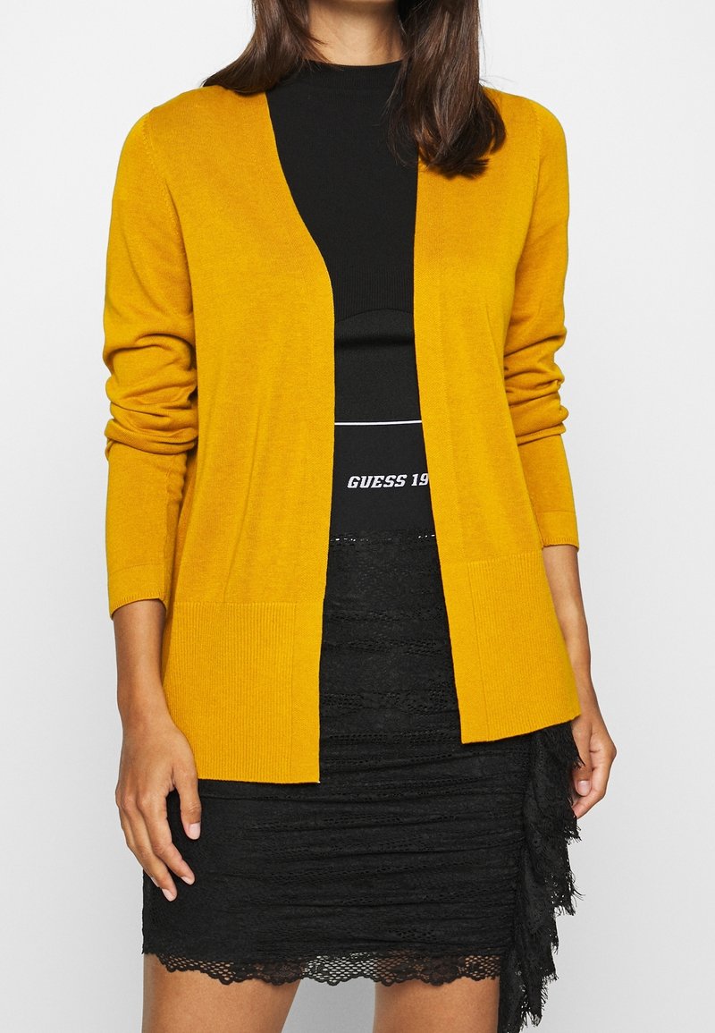 Cardigan en tricot jaune à manches longues et à l'avant ouvert, associé à un haut noir ajusté et à une jupe en dentelle noire texturée.