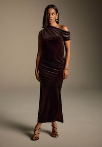 Brūns samta kleita ar vienu plecu, rušinājumu, sānu šķiršanu un potīšu siksniņām. Aksesuāri ietver izteiksmīgas zelta auskarus un gludu frizūru.