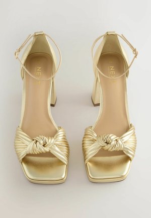 Zlaté vysoké sandály na podpatku s uzlovým, skladaným designem pásku, čtveratou špičkou a řemínkem kolem kotníku. Lesklý kovový povrch zvýrazňuje vizuální atraktivitu.