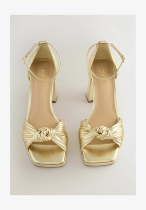 Kullanılmış altın rengi yüksek topuklu sandaletler, düğümlü ve plise tasarımlı kayış, kare burun ve bilek kayışıyla. Parlak metalik kaplama görsel çekiciliği artırıyor.