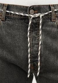 Κοντινή λήψη μαύρου τζιν τζιν παντελονιού που στερεώνεται με μια μαυρο-λευκή κορδέλα που περνάει μέσα από τις θηλιές της ζώνης και δένεται σε φιόγκο στη μέση.