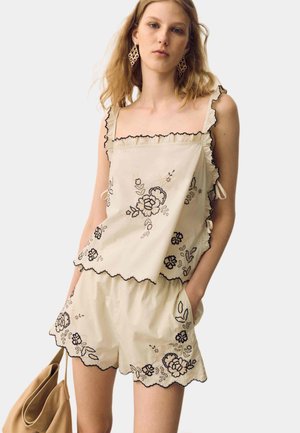 Ung kvinde iført beige blomsterbroderet ærmeløs top og matchende shorts, der holder en tanfarvet håndtaske og poserer mod en ensfarvet baggrund.