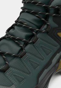 Groene wandelschoen met gestructureerde stof, zwarte veters en gele accenten. Voorzien van opvallende groeven en een versterkte neuskap.