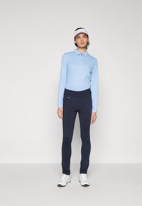Helles blaues Langarmpolo mit Reißverschlusskragen, kombiniert mit marineblauen, taillierten Hosen und weißen Sportschuhen. Die Schirmmütze ist weiß.