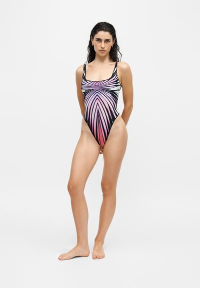 Femme aux cheveux foncés portant un maillot de bain une pièce avec un motif abstrait rayé rose, violet et noir, debout pieds nus sur fond blanc.