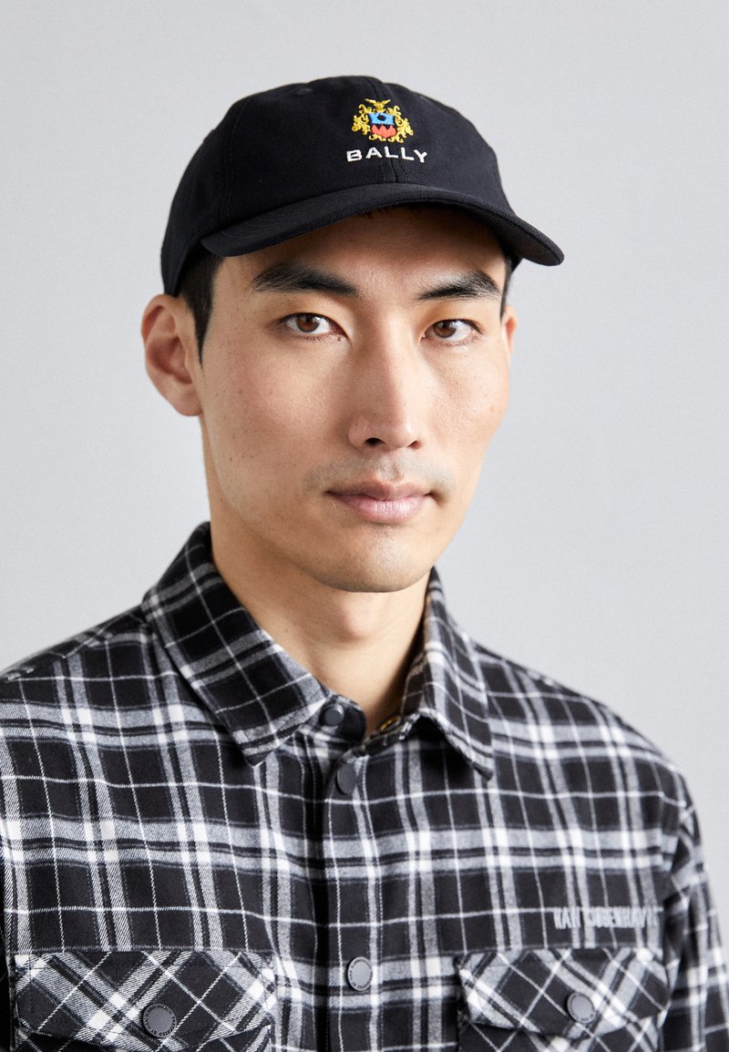 Black cap with yellow and blue emblem, white "BALLY" text; model wears a black and gray plaid flannel shirt with button pockets.