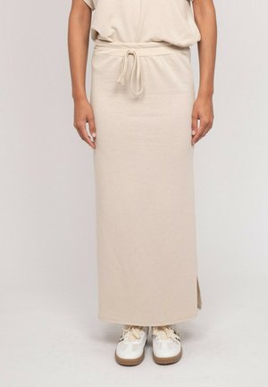 Personne portant une jupe beige à cordon, associée à des baskets blanches et beiges, debout devant un fond clair uni.