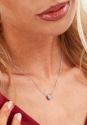 Silberne Kettennocke mit einem runden, klaren Edelstein-Anhänger. Der Hintergrund zeigt Hauttöne und Haare.