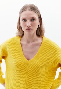 Geel gebreide trui met een v-hals, voorzien van ribbelstructuur en lange mouwen. Het materiaal lijkt zacht en behaaglijk.