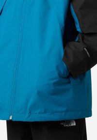 Blauwe waterdichte jas met zwarte accenten, ritssluiting aan de voorkant en logo op de zoom, met een gladde textuur en losse pasvorm.