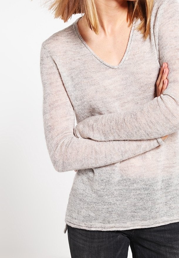 Femme portant un pull léger à manches longues couleur beige clair avec un col en V, les bras croisés, associé à un pantalon foncé, sur un fond blanc.