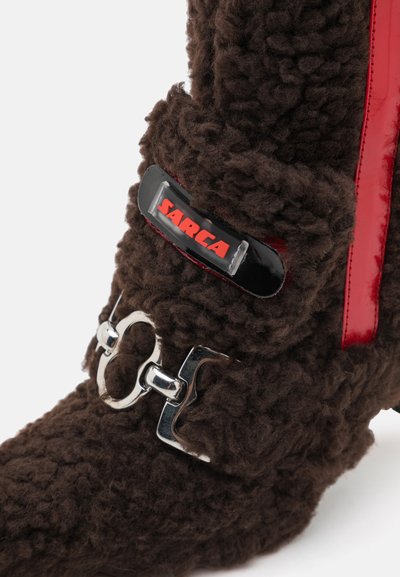 Bota marrón de textura esponjosa con un acento de cuero patente rojo, herrajes plateados y una etiqueta negra prominente con texto rojo.