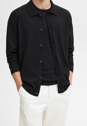 Homme portant une chemise noire à manches longues boutonnée par-dessus un t-shirt noir, avec un pantalon blanc et les mains dans les poches, sur un fond uni.