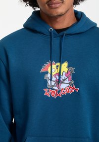 Blå hættetrøje med et grafisk print af to fugle og en globe, samt et rødt "VOLCOM" logo. Materialet ser blødt ud, med en afslappet pasform og snoretræk i hætten.