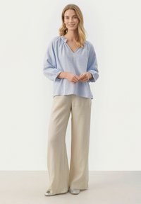 Lys blå linned blouse med V-hals og rynkedetalje langs udskæringen, parret med cremede, vide linnedbukser og sølvfarvede sko.