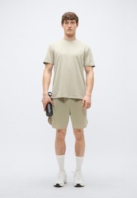 Conjunto atlético cáqui claro composto por uma t-shirt de manga curta e calções. Usado com meias brancas até ao meio da canela e ténis, segurando uma garrafa de água preta.