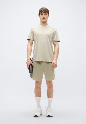 Αθλητική ενδυμασία σε ανοιχτό χακί χρώμα που περιλαμβάνει κοντομάνικη μπλούζα και σορτς. Φοριέται με λευκές κάλτσες και αθλητικά παπούτσια, κρατώντας ένα μαύρο παγούρι νερού.