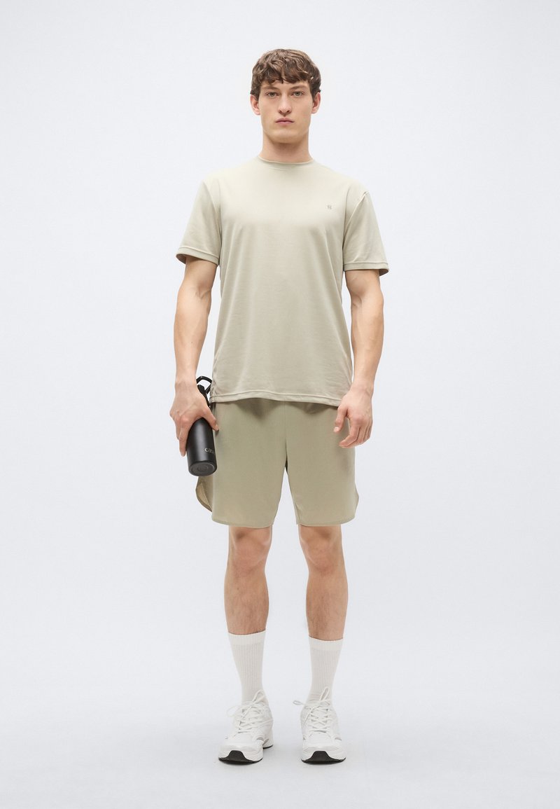 Conjunto atlético cáqui claro composto por uma t-shirt de manga curta e calções. Usado com meias brancas até ao meio da canela e ténis, segurando uma garrafa de água preta.