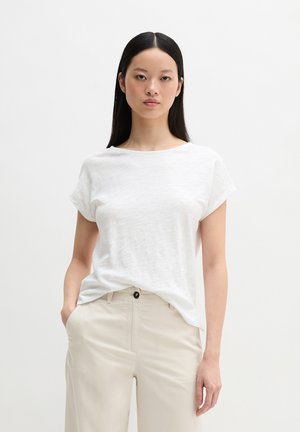 Frau mit langen schwarzen Haaren, die ein weißes Kurzarmoberteil und beige Hosen trägt, mit einer Hand in der Tasche vor einem einfarbigen Hintergrund stehend.