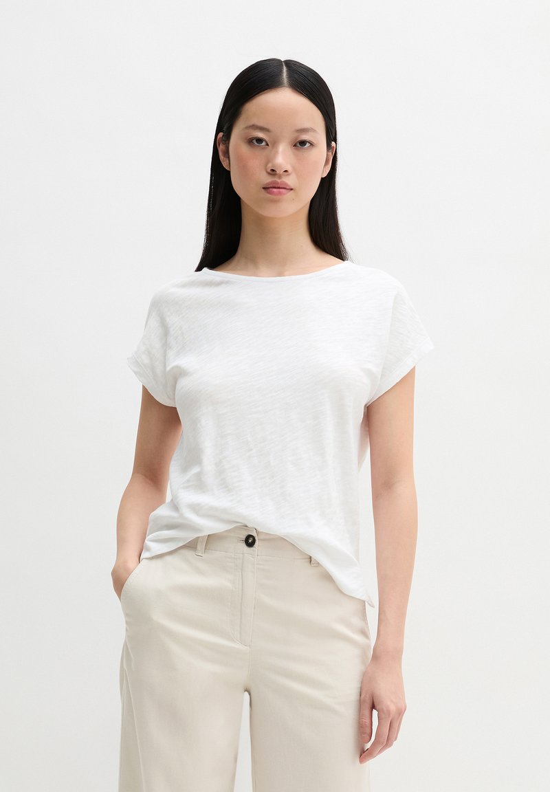 Γυναίκα με μακριά μαύρα μαλλιά που φοράει λευκό μπλουζάκι με κοντά μανίκια και μπεζ παντελόνι, στέκεται με το ένα χέρι στην τσέπη μπροστά σε απλό φόντο.