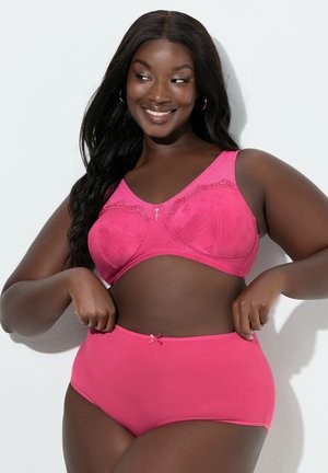 Lächende Frau passt hoch taillierte pinke Unterwäsche an, trägt dazu passenden Spitzen-BH und steht vor weißem Hintergrund.