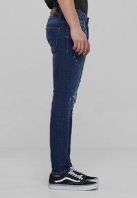 Jeans de mezclilla azul delgados con un acabado desgastado y detalles rasgados, combinados con zapatillas de corte bajo en blanco y negro, mostrados de lado.