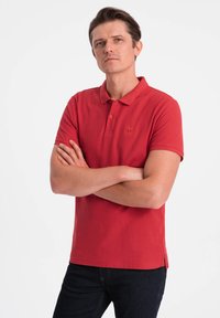 Polo rouge à manches courtes en coton, avec un col, trois boutons et un petit logo. Associé à un jean foncé.