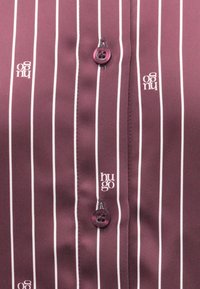Bordová košile na knoflíky s bílými svislými pruhy. Má lesklý povrch, fialové knoflíky a logo značky rozmístěné podle vzoru.